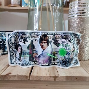 Peut inclure: Un billet d'un dollar modifié avec des personnages de Star Wars. La facture affiche Luke Skywalker, Dark Vador, la princesse Leia, Yoda et R2-D2. Le texte "FEDERAL RESERVE NOTE" et "ONE DOLLAR" sont visibles.