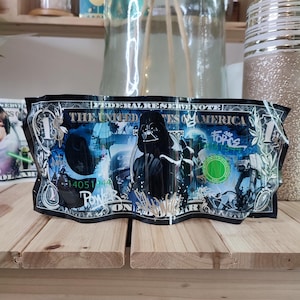 Peut inclure: Un billet d'un dollar avec un motif bleu et vert représentant Dark Vador de Star Wars. Le billet porte les mentions "Federal Reserve Note" et "The United States of America". Le billet est froissé et porte une signature.