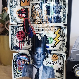 K&ouml;nnte beinhalten: Ein dreidimensionales Kunstwerk mit Dollarnoten mit Graffiti-Kunst und Portr&auml;ts. Das Kunstwerk enth&auml;lt Bilder eines Dinosauriers, einer Krone, eines Boxers und den Text "NEW YORK". Die untere Note zeigt ein Portr&auml;t von Jean-Michel Basquiat.