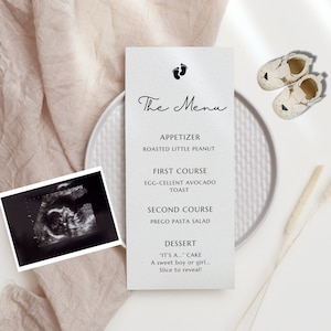 Puede incluir: Una tarjeta blanca con texto negro sobre un plato blanco con un mantel de color beige claro. El texto dice "The Menu" y enumera los elementos del menú: "Aperitivo: Roasted Little Peanut", "Primer plato: Egg-cellent Avocado Toast", "Segundo plato: Prego Pasta Salad" y "Postre: "It's A..." Cake. A sweet boy or girl... Slice to reveal!"