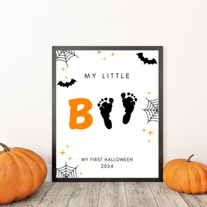 K&ouml;nnte beinhalten: Ein schwarzer gerahmter Druck mit wei&szlig;em Hintergrund und orangefarbenem und schwarzem Text. Der Text lautet "My Little Boo" mit einem Paar Baby-Fu&szlig;abdr&uuml;cken. Der Druck enth&auml;lt auch ein Spinnennetz, Flederm&auml;use und Sterne. Der Text unten lautet "My First Halloween 2024".