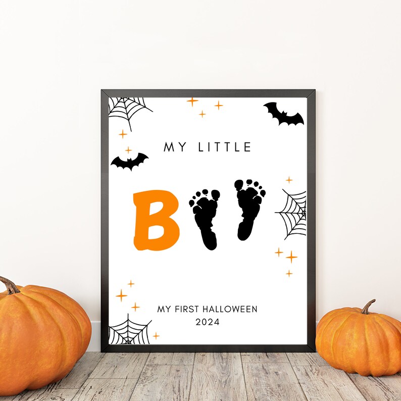 K&ouml;nnte beinhalten: Ein schwarzer gerahmter Druck mit wei&szlig;em Hintergrund und orangefarbenem und schwarzem Text. Der Text lautet "My Little Boo" mit einem Paar Baby-Fu&szlig;abdr&uuml;cken. Der Druck enth&auml;lt auch ein Spinnennetz, Flederm&auml;use und Sterne. Der Text unten lautet "My First Halloween 2024".