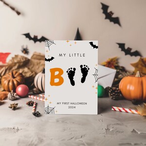 K&ouml;nnte beinhalten: Eine wei&szlig;e Karte mit orangefarbenem und schwarzem Text, der "My Little Boo" und "My First Halloween 2024" lautet. Die Karte hat schwarze Fu&szlig;abdr&uuml;cke und ein Spinnennetz-Design.
