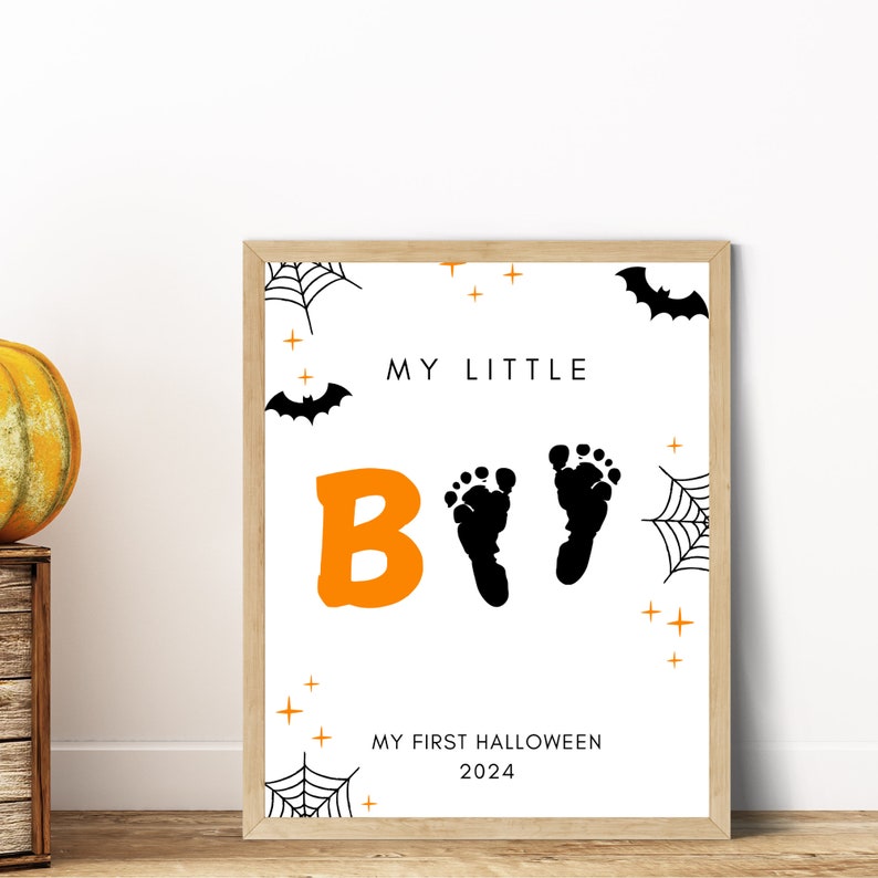 K&ouml;nnte beinhalten: Ein wei&szlig;es bedruckbares Schild mit orangefarbenem und schwarzem Text und Grafiken. Der Text lautet "My Little Boo" mit zwei schwarzen Fu&szlig;abdr&uuml;cken. Der Text unten lautet "My First Halloween 2024".