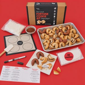May include: A Global Grub fortune cookie kit including a cardboard box, a baking mat, a spatula, a marker, a small bowl of cocoa powder, a bag of mix, a tray of baked fortune cookies, and a sheet of paper with fortune text prompts.