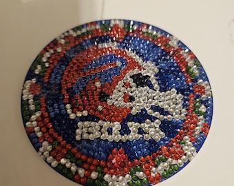May include: A circular coaster with a sparkling design in blue, red, white, and green. The coaster has the word "BILLS" in silver rhinestones, with a football helmet design. The coaster is likely made of a hard, durable material.
