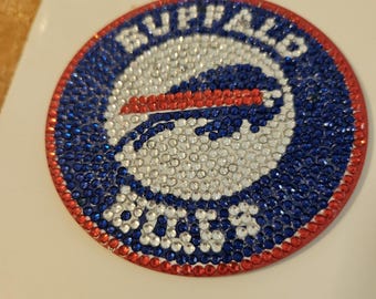 May include: A circular emblem featuring the Buffalo Bills logo, encrusted with sparkling rhinestones. The design includes a blue and red border, with the team name and logo in white and blue. The emblem is on a white surface.