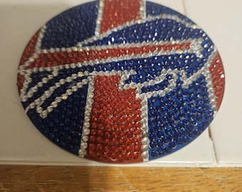 May include: A circular decorative piece with a design in red, white, and blue rhinestones. The design is a stylized logo or emblem, with a red section and a blue background. The rhinestones create a sparkling, textured effect, adding a touch of glamour.