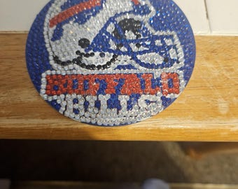 May include: A circular, blue, white, and red Buffalo Bills logo, embellished with rhinestones. The logo depicts a football helmet and the team name. The design is on a wooden surface.