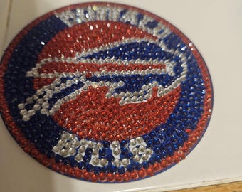 May include: A circular, bedazzled emblem featuring the Buffalo Bills logo. The design incorporates red, white, and blue rhinestones, with the team name and logo clearly visible. The emblem is set against a white background.