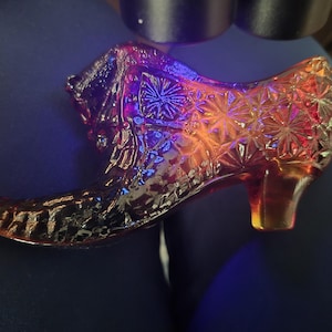 Op de afbeelding: Een decoratief glazen schoenbeeldje met een levendige iriserende afwerking die verschuift tussen dieppaarse, oranje en rode tinten. Het oppervlak is versierd met een gedetailleerd geperst sterren- en geometrisch patroon op de hiel en zijkant.