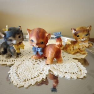 May include: Three ceramic kitten figurines in shades of gray, orange, and brown, each adorned with a bow. They are displayed on delicate white lace doilies, set on a silver tray.