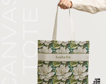 May include: Canvas tote bag with a floral pattern of white flowers and green leaves on a dark teal background. The name "Isabella" is printed on a light beige band across the front. The bag is held by a person.