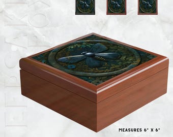 Puede incluir: Una caja de madera con un diseño de libélula. La caja es marrón con una parte superior tallada detallada que presenta una libélula azul rodeada de follaje verde. La caja mide 15 cm x 15 cm. Tres diseños cuadrados más pequeños con motivos de libélulas son visibles arriba.