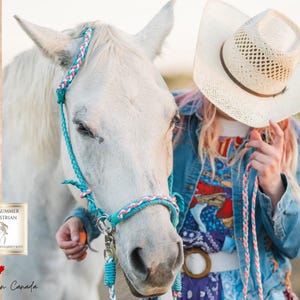Könnte beinhalten: Ein weißes Pferd mit einem türkisfarbenen und rosa geflochtenen Halfter. Eine Person mit einem Cowboyhut aus Stroh und einer Jeansjacke hält die Führungsleine. Das Bild enthält den Text "OPAL SUMMER EQUESTRIAN" und "Handmade in Canada".