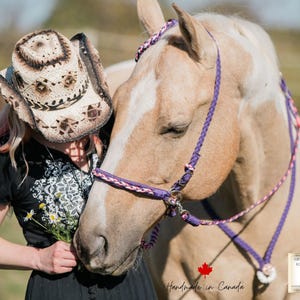 Könnte beinhalten: Ein hellbraunes Pferd mit einem geflochtenen Halfter in Lila und Rosa. Eine Person mit einem Cowboyhut aus Stroh lehnt sich an das Pferd. Der Hut hat ein dekoratives Muster. Das Bild ist im Freien aufgenommen.