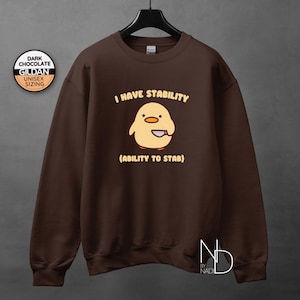 May include: Dark chocolate brown Gildan unisex sweatshirt with the text "I HAVE STABILITY (ABILITY TO STAB)" and a cartoon chick with a knife. The sweatshirt is hanging on a black hanger.