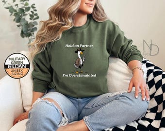 May include: Military green Gildan unisex sweatshirt with the text "Hold on Partner, I'm Overstimulated" and a cartoon goose wearing a hat and holding a gun. The sweatshirt is a solid color and has a round neckline.