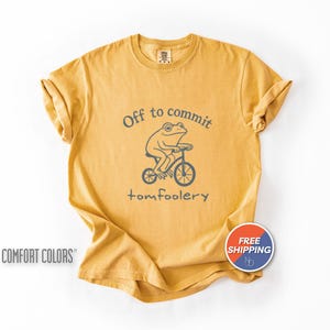 May include: Mustard yellow t-shirt with a frog riding a bicycle graphic. The text above the frog reads "Off to commit" and below the frog reads "tomfoolery". The shirt has rolled sleeves and a "Comfort Colors" logo.