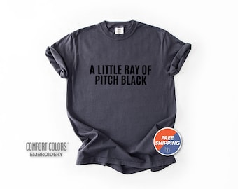 May include: A charcoal gray short-sleeve t-shirt featuring the text A LITTLE RAY OF PITCH BLACK printed in bold black letters across the chest. The garment has a crew neckline and a relaxed fit, with the Comfort Colors brand label visible at the neck.
