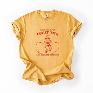 May include: Mustard yellow t-shirt with the text "SORRY FOR HAVING GREAT TITS AND CORRECT OPINIONS" in red. The graphic features a vintage-style illustration of a woman jumping rope. The shirt has rolled-up sleeves.