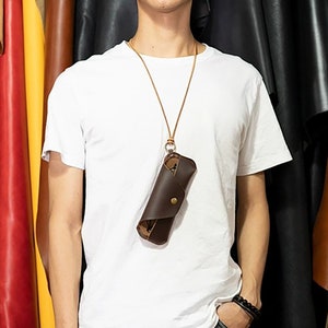May include: A brown leather glasses case with a button closure, suspended from a tan leather cord. The case is worn around the neck. The background features various colours of leather.