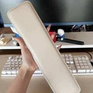 May include: A beige wrist rest for a keyboard. The wrist rest is made of a soft, padded material and has a stitched edge.