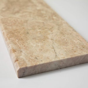 May include: A rectangular slab of beige marble with subtle veining and a polished surface. The stone has a light brown hue with darker, irregular patterns. The edge is a lighter, solid color. Ideal for countertops or decorative elements.