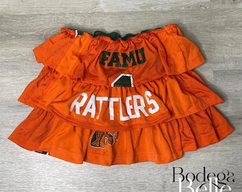 May include: An orange tiered skirt with the words "FAMU" and "RATTLES" in white lettering. The skirt has a mesh-like texture and a green trim at the top. The skirt appears to be made from a jersey material.