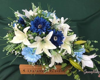 May include: A floral arrangement featuring white lilies, blue gerbera daisies, and accents of greenery. The arrangement is set on a wooden base with the engraved text "Groovy Ruby". The background is a dark green fabric.