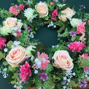 May include: A floral wreath featuring peach roses, pink carnations, white chrysanthemums, and small purple flowers. The wreath is surrounded by green foliage and small white flowers. The wreath is set against a dark green background.