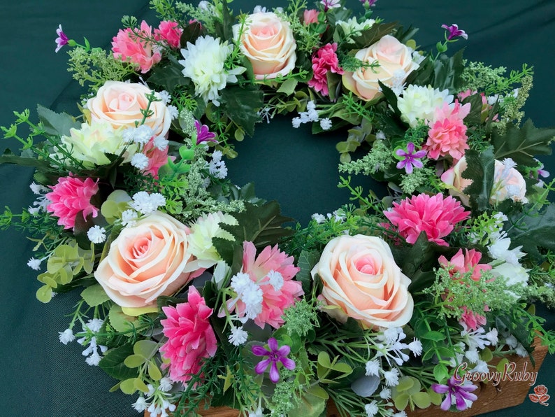 May include: A floral wreath featuring peach roses, pink carnations, white chrysanthemums, and small purple flowers. The wreath is surrounded by green foliage and small white flowers. The wreath is set against a dark green background.