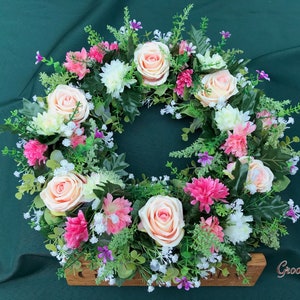 May include: A floral wreath featuring peach roses, pink carnations, white chrysanthemums, and small purple flowers. The wreath is set against a dark green background and includes greenery. The wooden base has the text "GroovyRuby".