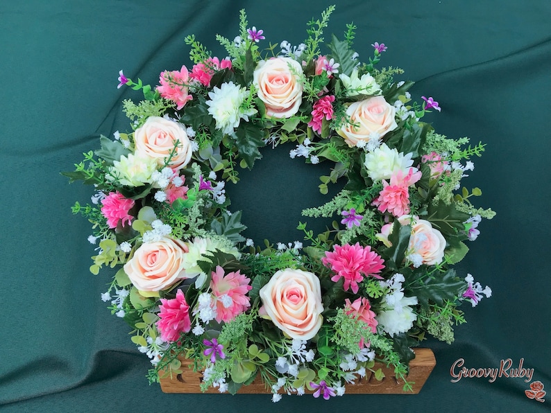May include: A floral wreath featuring peach roses, pink carnations, white chrysanthemums, and small purple flowers. The wreath is set against a dark green background and includes greenery. The wooden base has the text "GroovyRuby".