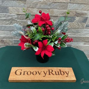 May include: A floral arrangement featuring vibrant red poinsettia flowers, green leaves, frosted pine branches, and small red berries in a black container, accompanied by a rectangular wooden plaque engraved with the text GroovyRuby.