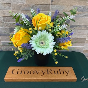 May include: An artificial flower arrangement in a black vase, featuring yellow roses, white gerbera daisies and purple lavender. The arrangement is displayed on a green surface with a wooden sign that reads "Groovy Ruby".