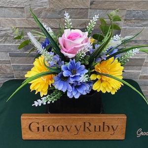 May include: A floral arrangement in a black vase with a pink rose, yellow and blue flowers, and green foliage. The arrangement is on a green surface with a wooden sign that reads "GroovyRuby".
