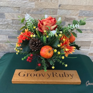 May include: An artificial flower arrangement in a brown vase, featuring orange roses, red berries, orange and yellow flowers, and a pine cone. The arrangement is on a green surface with a wooden sign that reads "Groovy Ruby".