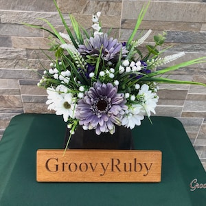 May include: A bouquet of white and purple flowers in a black vase with a wooden sign that reads "GroovyRuby".