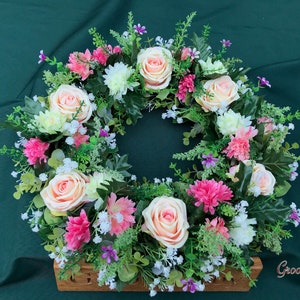May include: A floral wreath featuring peach roses, pink carnations, white daisies, and small purple flowers. The wreath is set in a wooden base and surrounded by green foliage. The GroovyRuby logo is visible in the bottom right corner.