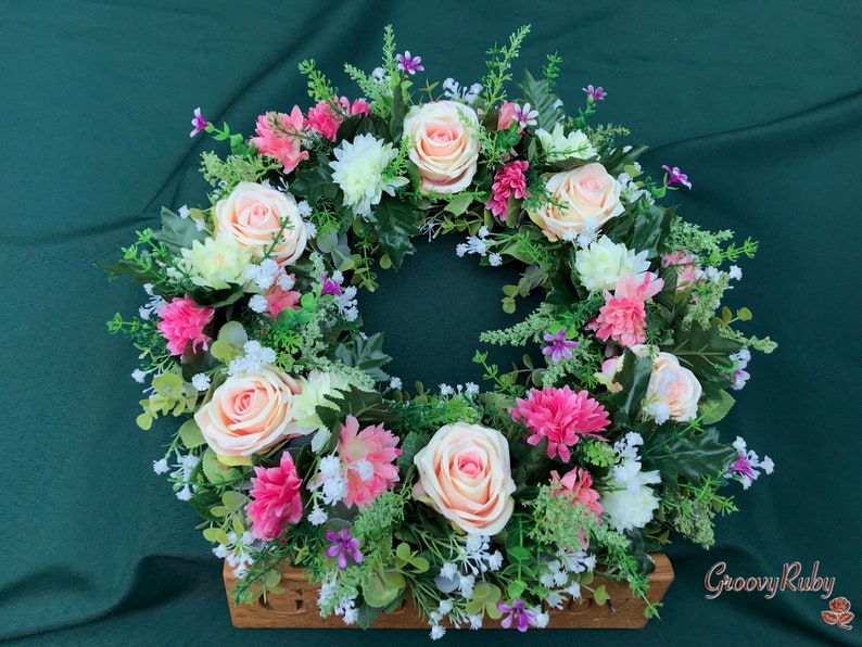 May include: A floral wreath featuring peach roses, pink carnations, white daisies, and small purple flowers. The wreath is set in a wooden base and surrounded by green foliage. The GroovyRuby logo is visible in the bottom right corner.