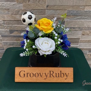 May include: An artificial flower arrangement in a black vase with a football on top. The arrangement features white, yellow and blue flowers and is on a green surface with a wooden sign that reads "GroovyRuby".