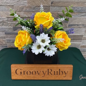 May include: A yellow and white artificial flower arrangement in a black square vase with a wooden sign that reads "GroovyRuby".