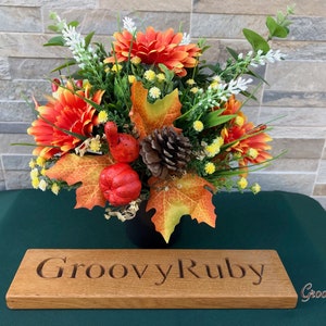 May include: A fall-themed floral arrangement in a black pot with orange and yellow flowers, green leaves, and pine cones. The arrangement is sitting on a green surface with a wooden sign that reads "Groovy Ruby".