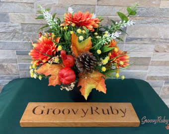 May include: A fall-themed floral arrangement in a black pot with orange and yellow flowers, green leaves, and pine cones. The arrangement is sitting on a green surface with a wooden sign that reads "Groovy Ruby".