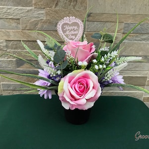 May include: A pink and purple artificial flower arrangement in a black pot with a heart-shaped sign that says "Happy Mother's Day".