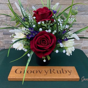 May include: A red and white artificial flower arrangement in a black pot with a wooden sign that reads "Groovy Ruby".