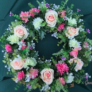 May include: A circular floral wreath featuring peach roses, pink and white chrysanthemums, and small purple flowers. Green foliage and sprigs of white flowers fill the gaps between the blooms. The wreath is set against a dark green background.