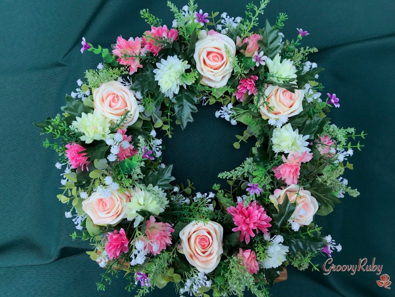 May include: A circular floral wreath featuring peach roses, pink and white chrysanthemums, and small purple flowers. Green foliage and sprigs of white flowers fill the gaps between the blooms. The wreath is set against a dark green background.