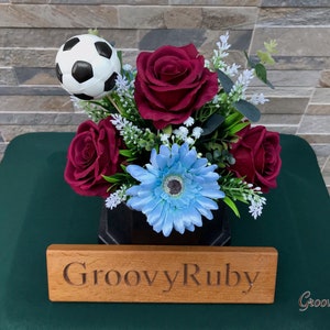 May include: A black and white football sits in a bouquet of red roses and a light blue gerbera daisy. The flowers are in a black vase with a wooden sign that reads "GroovyRuby".